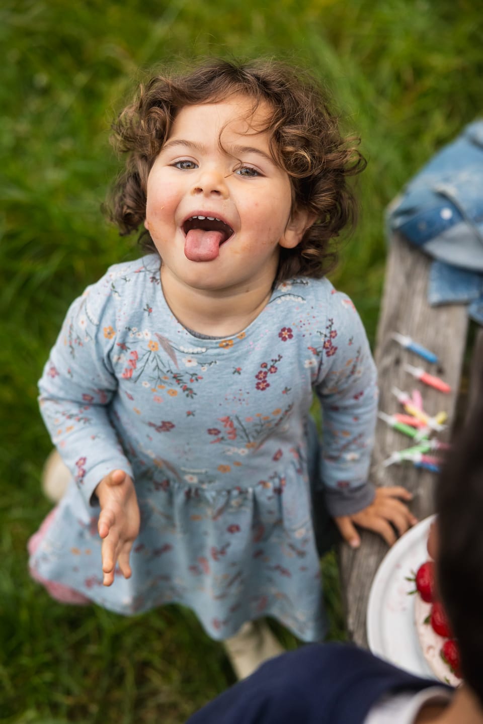 A laughing toddler with her tongue out reaching forward in a garden.