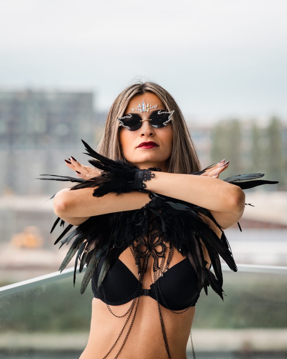 A studio-lit portrait of a woman in a black feather harness with her arms crossed.