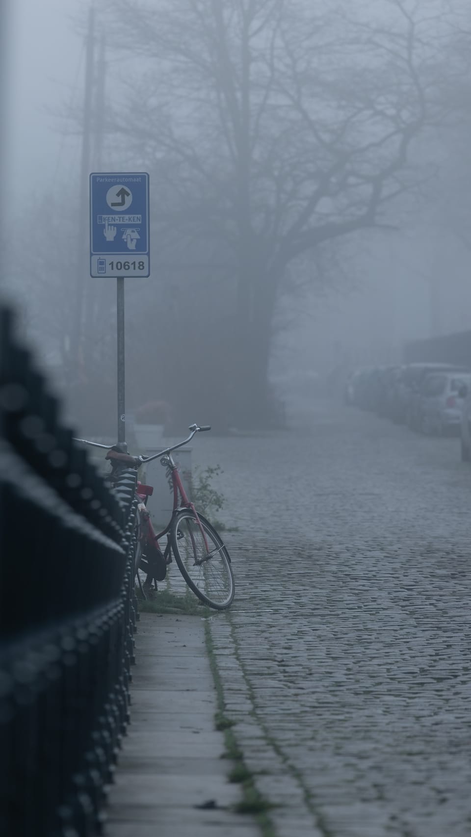 A lone bicycle leaning on a foggy cobblestone quay lined with bare trees.