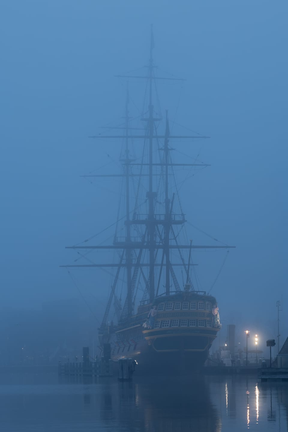 The masts of a tall ship rising through blue-hour fog, lights reflecting on the water.