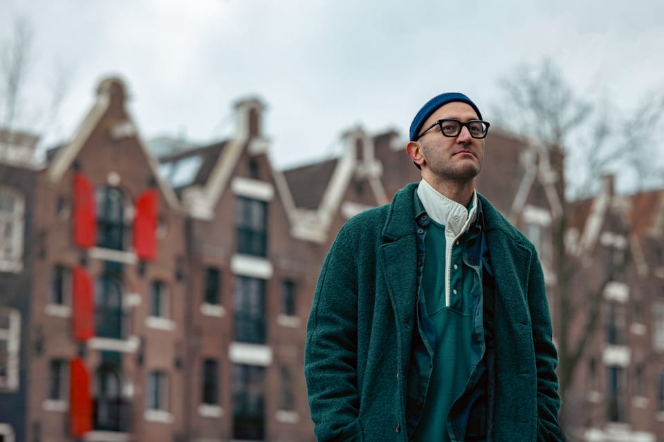 A man in a green coat and beanie standing before Amsterdam canal houses.