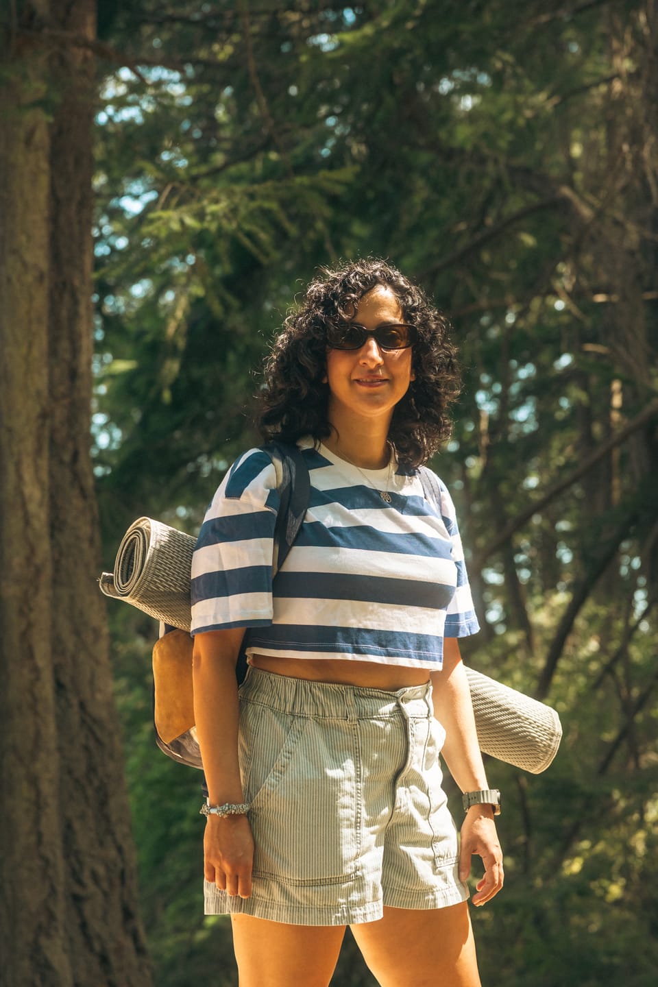 A woman with curly hair and a striped shirt standing in a sunlit forest.