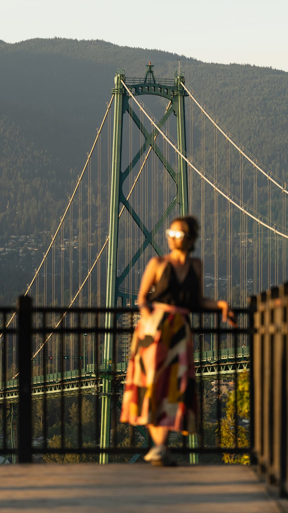A woman on a walkway with the Lions Gate Bridge rising behind her at golden hour.