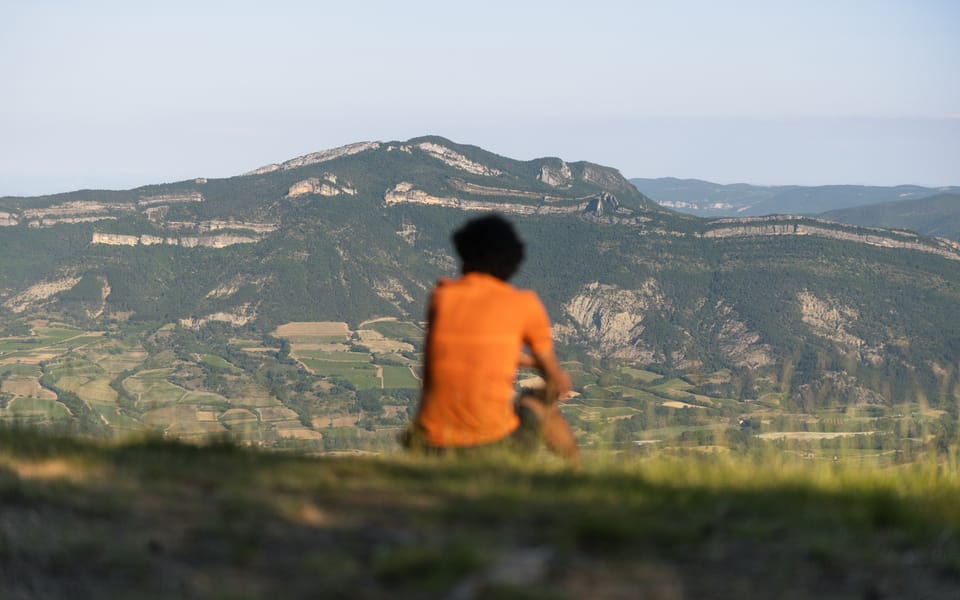 A person in an orange shirt sitting on a hilltop looking out over distant mountains.