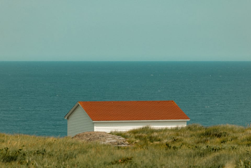 A small red-roofed hut standing alone on a green headland above a teal sea.