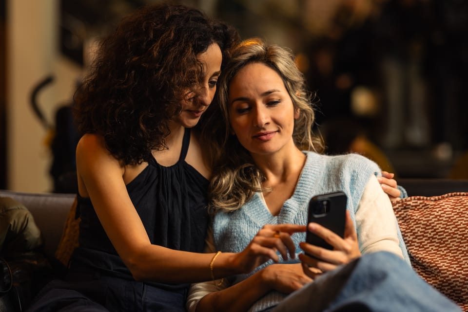 Two women leaning together on a sofa, looking at a phone in warm light.