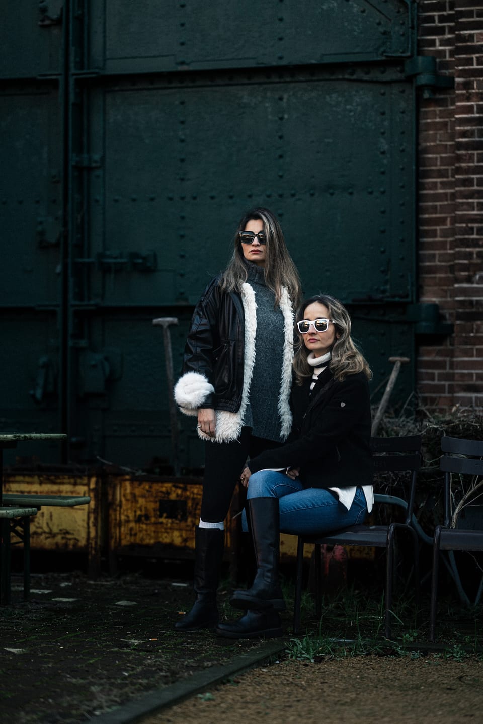 Two women in fur-trimmed jackets and sunglasses seated against a green industrial door.