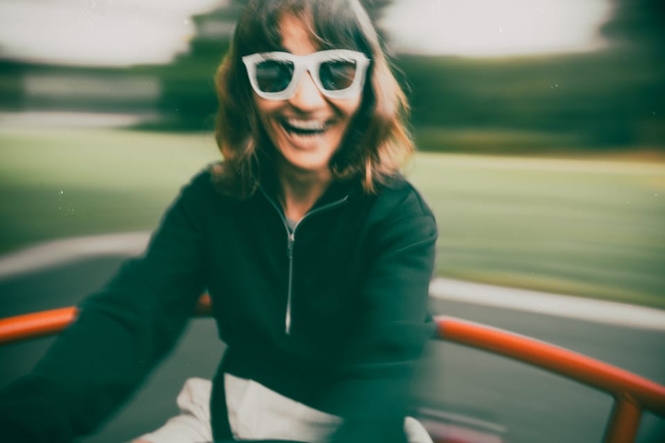 A woman in white sunglasses laughing in a moving convertible, the world blurred behind her.
